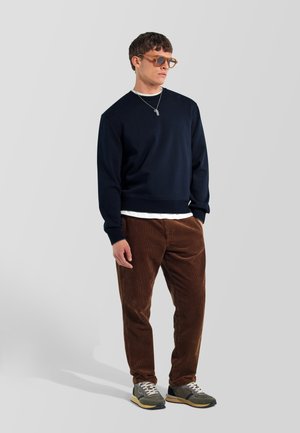 Uomo che indossa pantaloni di velluto a coste marroni, felpa blu scuro sopra camicia bianca, sneakers grigie, occhiali da sole e una collana, in piedi su uno sfondo semplice.