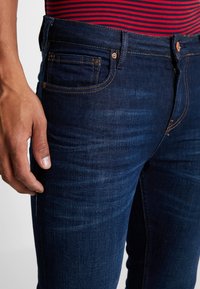 Vaqueros de mezclilla azul oscuro con un corte ajustado, costuras visibles y un bolsillo delantero. El material muestra una ligera textura con detalles sutiles de desgaste.