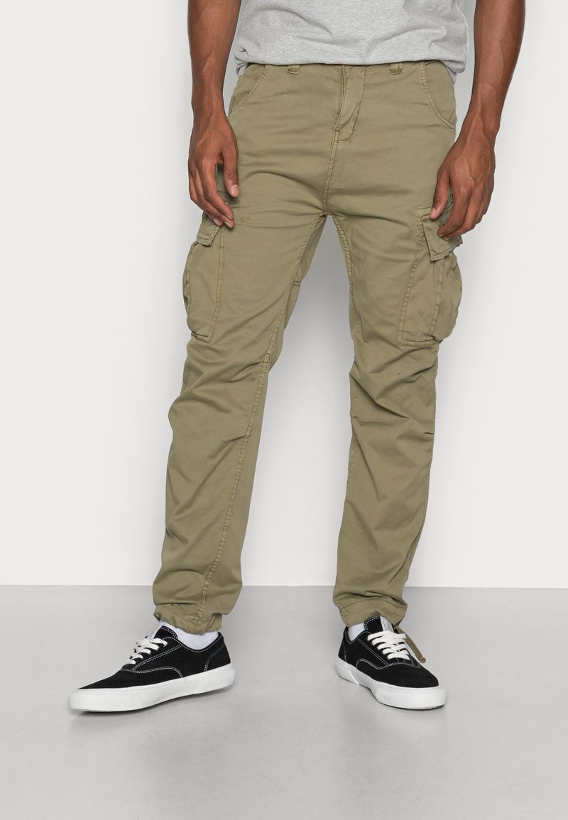 Homme portant un pantalon cargo beige et des baskets noires et blanches, debout sur un fond blanc.