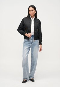 Μαύρο μπουφάν bomber με χρυσά φερμουάρ, ριμπ μανσέτες και λεία υφή, συνδυασμένο με ανοιχτό μπλε τζιν παντελόνι φαρδιάς γραμμής και μαύρες μπαλαρίνες.