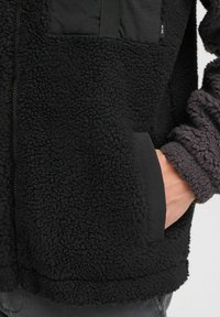 Chaqueta de forro polar negra con un exterior texturizado, cierre con cremallera y un bolsillo de tela contrastante. Los puños son suaves y ajustados.