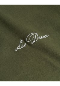 Olivgrüner Stoff mit glatter Textur, mit weißem, gesticktem Text "Les Deux" in einer kursive Schriftart. Keine zusätzlichen Details sichtbar.