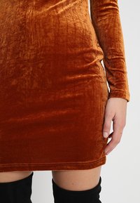 Samtkleid in einem satten, verbrannten Orangeton, figurbetont mit langen Ärmeln, glatter Textur und einem Saum kurz über dem Knie, kombiniert mit schwarzen Überknie-Stiefeln.