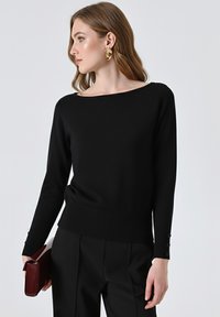 Maglione nero in maglia con scollatura a barca, maniche lunghe e dettagli con bottoni sui polsini, abbinato a pantaloni neri sartoriali e una borsa a tracolla bordeaux.
