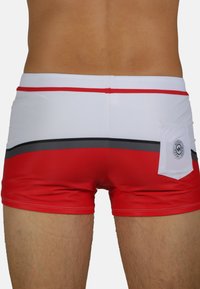 Un short de bain rouge avec un design rayé blanc et gris, comportant une petite poche arrière et une ceinture contrastante rouge. Matière lisse et extensible.