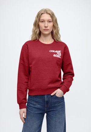 Une femme porte un sweat-shirt rouge à col rond avec un petit motif blanc sur la poitrine et un jean bleu sur un fond clair uni.
