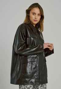 Must mustnahkjope, millel on tekstuuriga krokodillimuster, üle suurus, nööbitav krae ja kaks esikotti.