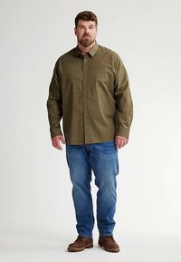 Grün gemustertes Hemd mit Knöpfen und langen Ärmeln, getragen über blauen Jeans und braunen Stiefeln. Das Hemd hat einen Kragen und vordere Knöpfe.