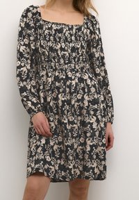 Kukallinen mekko, jossa on neliön muotoinen pääntie, pitkät hihat ja ryppydetaljit tummalla taustalla, jossa on kerma- ja hienovaraisia persikkasävyjä.