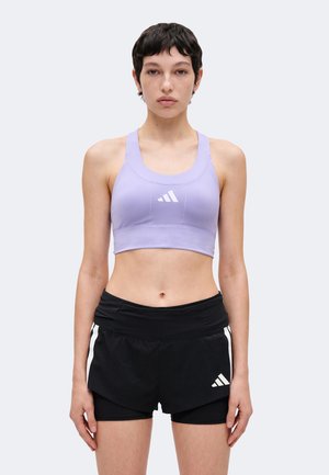 Jeune personne aux cheveux courts portant un brassière de sport lavande et un short de sport noir avec des logos Adidas blancs, debout face à l'avant.
