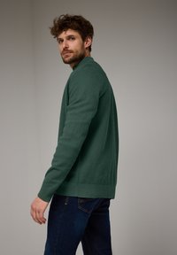 Pullover verde a maglia testurizzata con colletto alto e polsini a costine, abbinato a jeans blu scuro, mostrato di lato su uno sfondo neutro.