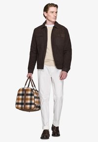 Brun ruskindsjakke over en cremefarvet kabelstrikket sweater, parret med hvide bukser. Holder en ternet duffelbag i nuancer af brun og sort.