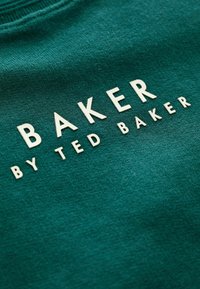 Zelená bavlnená tkanina s jemnou textúrou, na ktorej sú v tučných bielych písmenách napísané slová "BAKER" a nižšie menším písmom "by TED BAKER".
