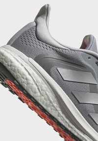Šedá sportovní bota s texturovaným síťovinovým svrškem, třemi překryvnými pruhy, bílou mezipodešví a červenou gumovou podrážkou s protiskluzovými vzory.
