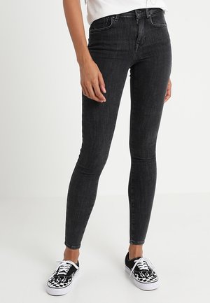 Person trägt schwarze Skinny Jeans und schwarz-weiße Sneakers mit Flammenmuster und steht vor einem schlichten Hintergrund.