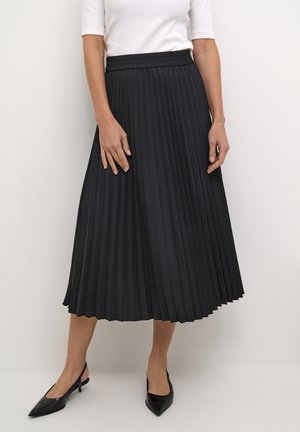 Femme portant une jupe midi plissée noire, un haut blanc à manches courtes et des escarpins slingback noirs à bout pointu, debout sur fond blanc.