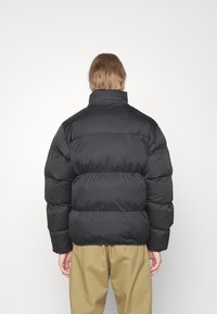 Sort pufferjakke med høj krave, quiltet design og blød tekstur, parret med brune, løstsiddende bukser.