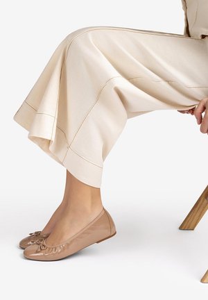 Legs crossed at the ankles wearing cream wide-leg pants and shiny nude ballet flats with small bows, seated on a wooden chair.