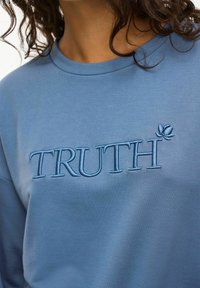 Ljusblå sweatshirt med rundad halsringning. Har det broderade ordet "SANNING" i en mörkare blå nyans. Mjuk tyg med en slät struktur.