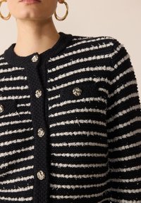 Randig kofta i svart och vitt med texturstickat material, silverdetaljer på knapparna och två framfickor.