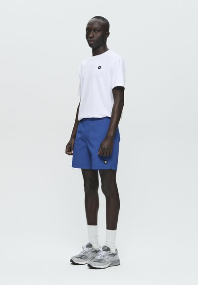Weißes T-Shirt mit kleinem Logo, blaue Shorts und graue Sneakers mit weißen Socken, die ein lässiges, sportliches Design zeigen.