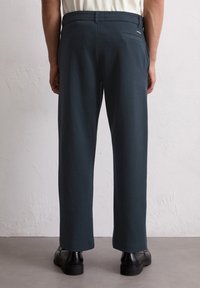 Pantalons vert foncé à coupe décontractée, fabriqués en tissu texturé. Comprend une poche arrière, portés avec des chaussures noires, montrant des coutures et des ourlets soignés.