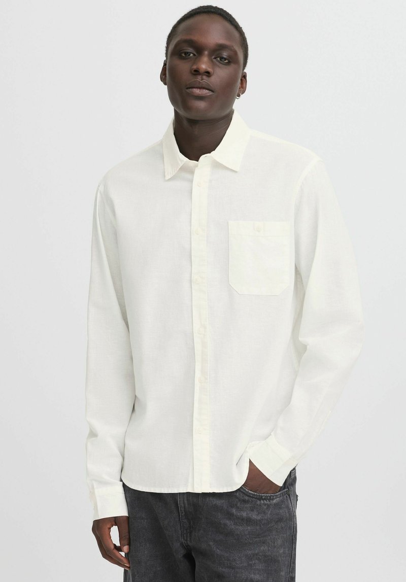 White long-sleeve shirt with a button-down collar and a chest pocket, made of a lightweight fabric with a slight texture. Worn with dark jeans.