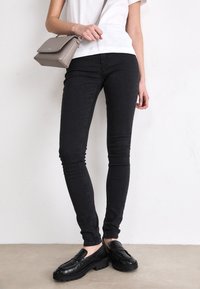 Zwarte skinny jeans met een gladde textuur, gecombineerd met zwarte instappers met een gestructureerd oppervlak, en een grijze schoudertas.