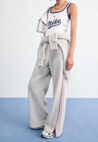 Person som bär en vit Nike tanktop, grå vida sweatpants med röda ränder, en knuten sweatshirt, guldhalsband, silverväska och sneakers.