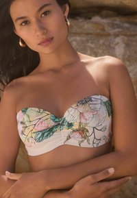 Bikini gornji dio s cvjetnim uzorkom u pastelnim bojama, nabranim središnjim šavom i mekim, glatkim materijalom. Zlatne naušnice u obliku obruča upotpunjuju izgled.