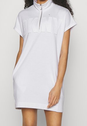 Kvinde iført en hvid kortærmet kjole med lynlåsforside og præget DKNY-logo over brystet.