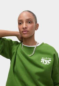 Felpa verde con vestibilità rilassata, presenta il testo ricamato bianco "NYC 1984" e una collana in argento, materiale in cotone, texture liscia.