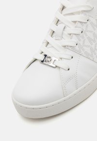 Hvit lær sneakers med strukturert sidepanel, tradisjonell snøring og et sølvfarget MK-logoaksent på snøringene. Myk gummisåle.