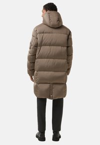 Bruine pufferjas met een capuchon, voorzien van gestikte secties, knoopsluiting aan de achterkant en een gladde textuur, gecombineerd met donkere broek en laarzen.