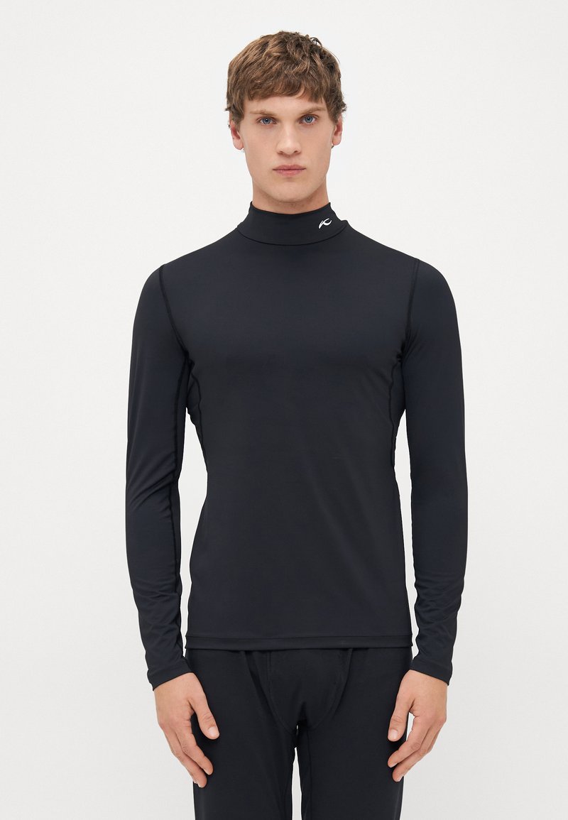 Young man wearing a fitted black long-sleeve mock neck top and black pants, standing against a plain white background.