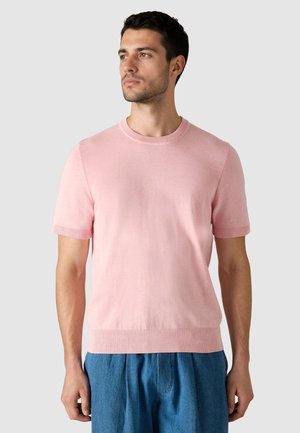 Mand iført en lyserød kortærmet striktrøje og løstsiddende blå bukser, der ser til venstre mod en ensfarvet lys baggrund.