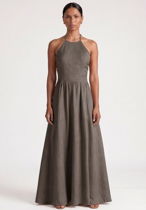 Kobieta ubrana w bezrękawową, oliwkowozieloną długą sukienkę z wiązaniem na szyi stojąca na tle jednolitego jasnego tła.