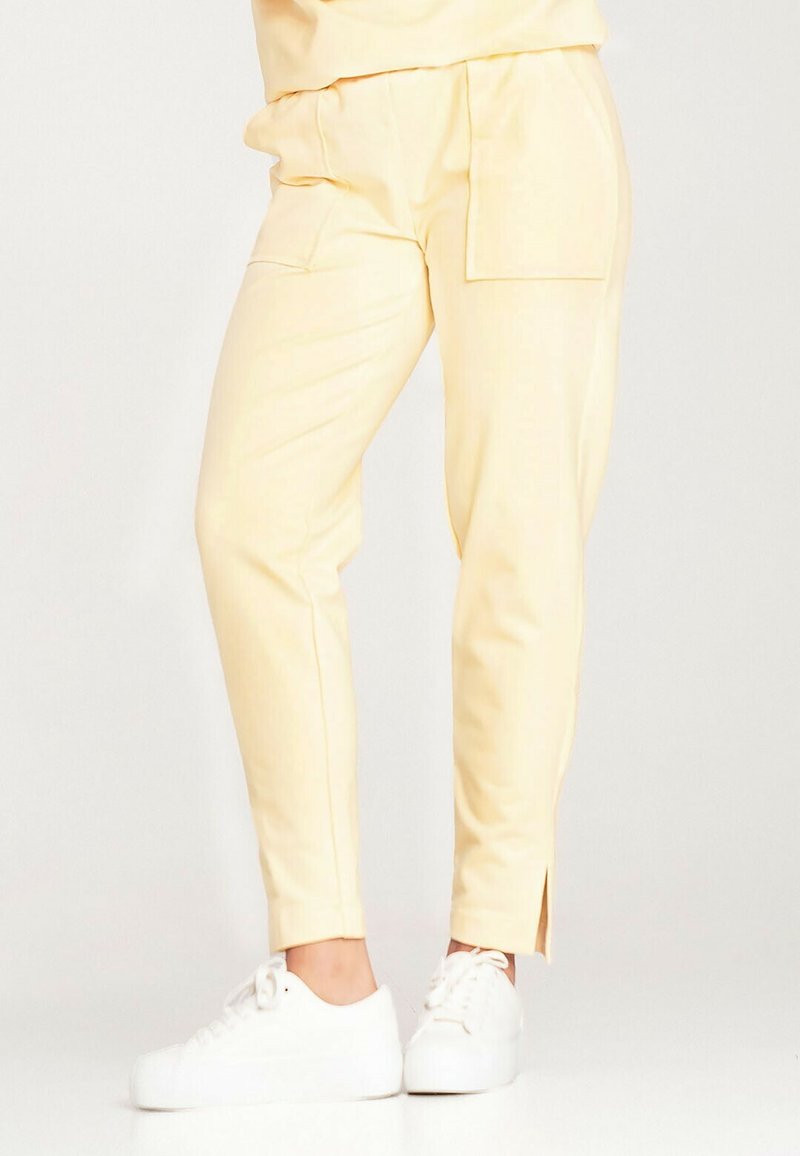 Żółte, zwężane spodnie wykonane z gładkiego materiału z bocznymi kieszeniami i rozcięciami na kostkach, w połączeniu z białymi sneakersami.