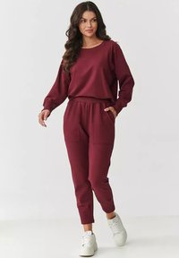 Burgundowa bluza i pasujące dresowe spodnie wykonane z miękkiego materiału, z elastycznym pasem, bocznymi kieszeniami i długimi rękawami. W zestawieniu z białymi sneakersami.