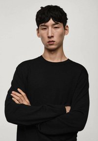 Maglione nero lavorato a maglia con collo rotondo, maniche lunghe e design aderente, indossato da una persona con le braccia incrociate su uno sfondo neutro.