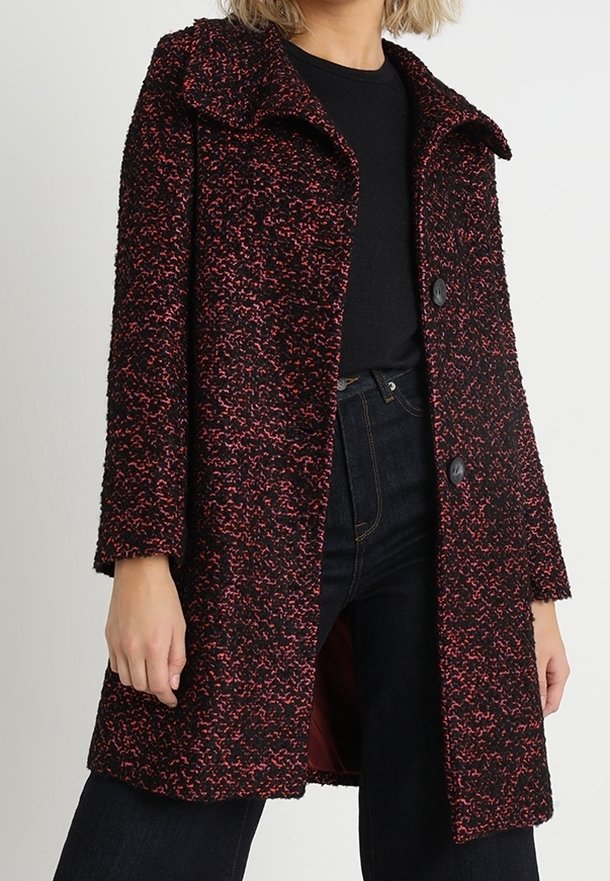 Femme portant un manteau texturé noir et rouge mi-long sur un haut noir et un jean foncé, debout avec les mains détendues le long du corps.