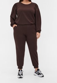 Brauner Pullover und passende Jogginghose aus weichem Material, mit puffierten Ärmeln und elastischen Bündchen, kombiniert mit schwarz-weißen Sneakers.