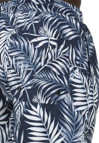Mörkblå badshorts med ett vitt mönster av palmlöv, en elastisk midjeband och en bakficka med svart tryckknappslåsning.