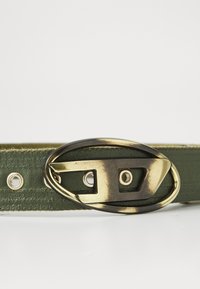 Ceinture en cuir vert olive avec une grande boucle ovales en bronze présentant un design ajouré et des œillets en métal pour l'ajustement.