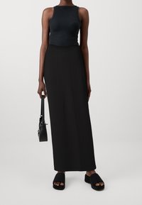 Schwarzes geripptes Top, langer schwarzer Rock und schwarze Sandalen mit offenen Zehen und Plattform. Schwarze Handtasche seitlich gehalten, durchgehend glatte Texturen.