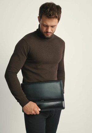 Czarna skórzana torba na laptopa trzymana przez osobę, o gładkiej fakturze i eleganckim designie, z minimalnymi zdobieniami i prostokątnym kształtem.