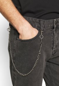 Jeans de mezclilla negra con una cadena de plata unida al bolsillo delantero, que presenta un detalle de broche y lazo. El material tiene una textura desgastada.