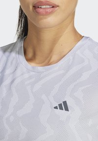 Primer plano de una persona que lleva una camiseta deportiva texturizada de color gris claro con un logo negro de Adidas en el pecho.