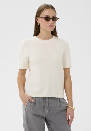 Suéter blanco de manga corta, con textura suave, cuello y dobladillo acanalados. Combinado con pantalones grises; el modelo lleva gafas de sol marrones y una pulsera de oro.