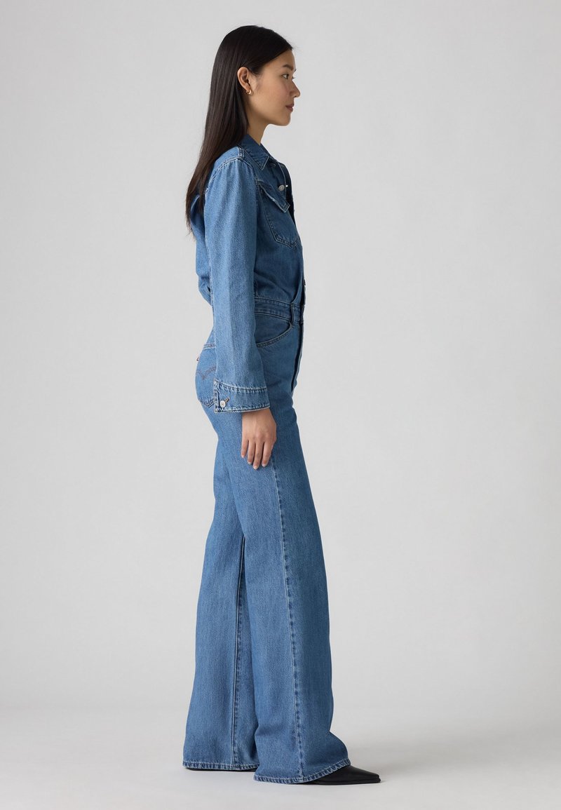 Combinaison en denim avec un haut ajusté à manches longues, taille haute et pantalon à jambes larges. Le matériau est un denim texturé bleu moyen.